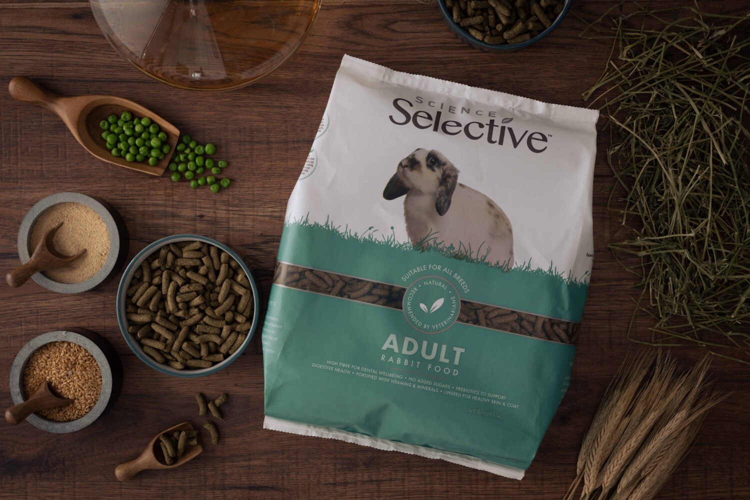 A bag of Science Selective Adult Rabbit Food is placed on a wooden surface, surrounded by natural ingredients such as green peas, hay, grains, and pellets. The packaging features a white and green design with an image of a lop-eared rabbit. Small bowls and wooden scoops hold various ingredients, emphasizing the food's natural composition.